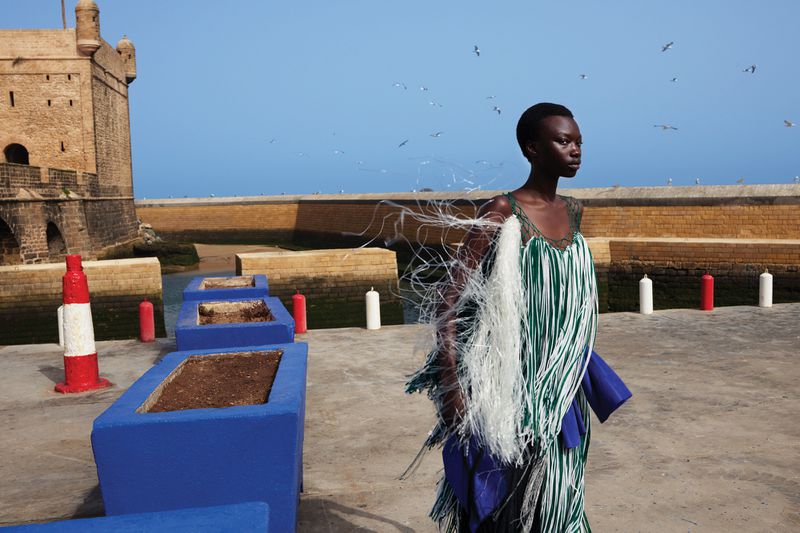 A slender woman stands in a flowing fringe dress against a blue sky and a stone fortress backdrop on a sunny day.
