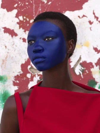 Headshot of dark-skinned female with short hair, blue painted face, wearing a red dress against a textured, patterned white wall.