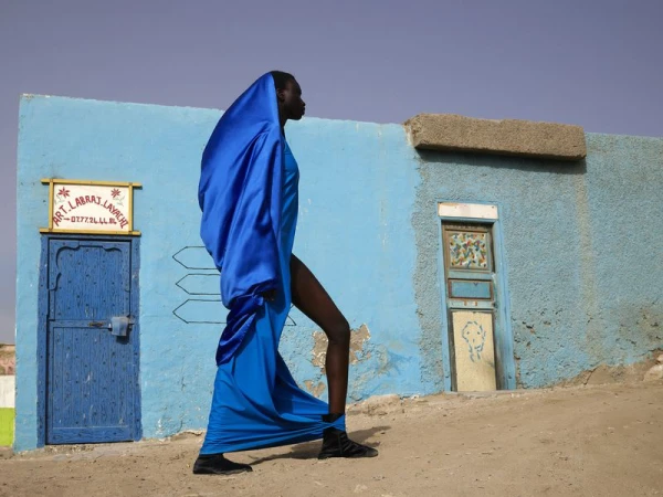 Fashion shot of a Black woman wearing a long blue silk dress, walking in front of a blue building with a door and a small window.