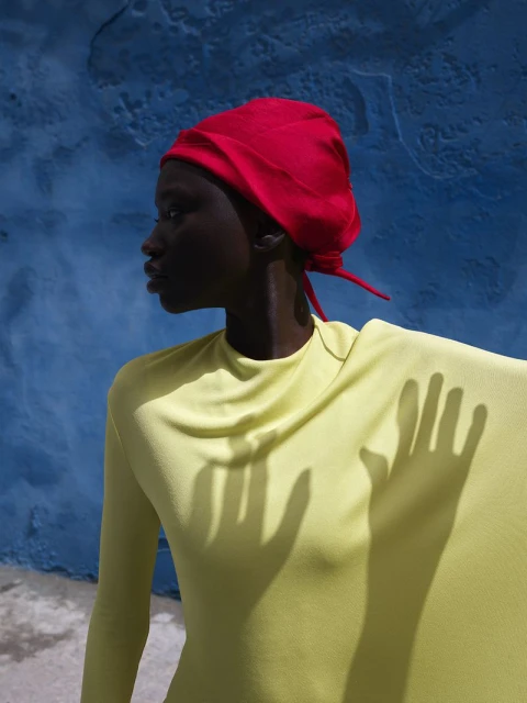 Profile shot of a woman with dark skin wearing a bright red headscarf and yellow top, with hand shadow on the garment against a blue wall.