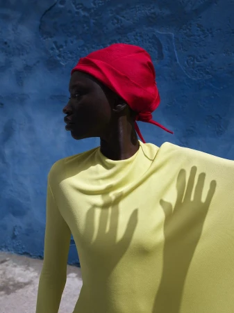 Profile shot of a woman with dark skin wearing a bright red headscarf and yellow top, with hand shadow on the garment against a blue wall.