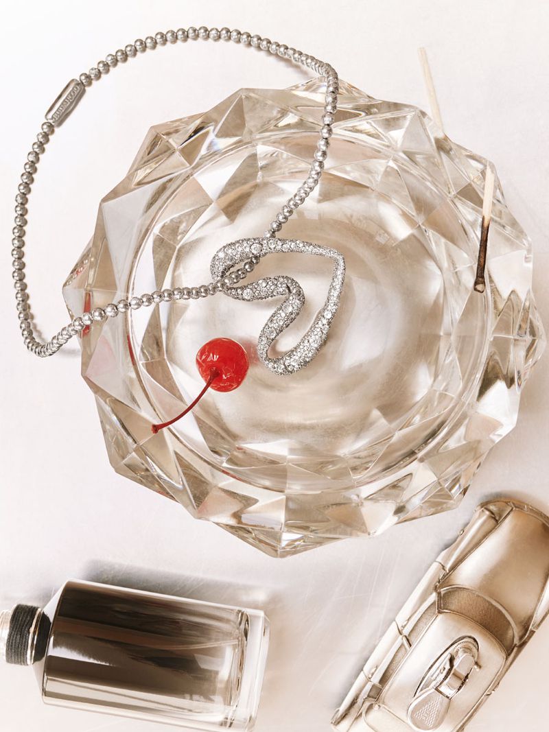 Close-up still life of a crystal ashtray holding a heart-shaped diamond necklace with a cherry, plus perfume and car model.