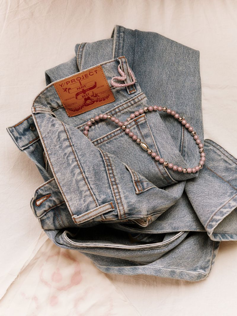 Folded light blue denim jeans and a beaded necklace arranged on a textured, pale pink surface, showcasing fashion and texture.