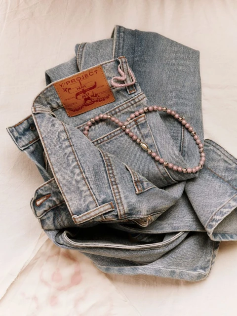 Folded light blue denim jeans and a beaded necklace arranged on a textured, pale pink surface, showcasing fashion and texture.