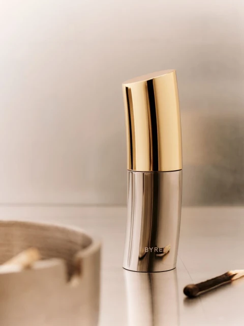 Still life of a Byredo fragrance bottle with a gold cap next to an ashtray with cigarette butts and a burnt matchstick on a neutral surface.