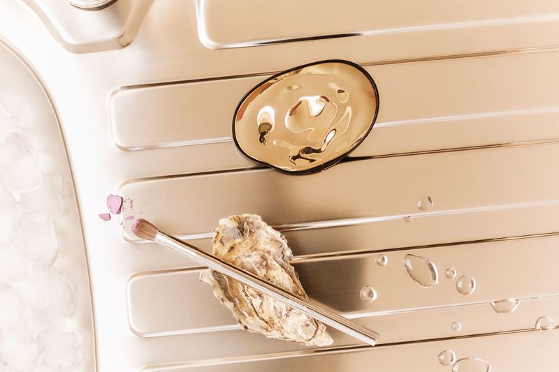 Still life shot of makeup brush, oyster shell, gold dish, water droplets, and pink powder arranged on a textured metallic background.
