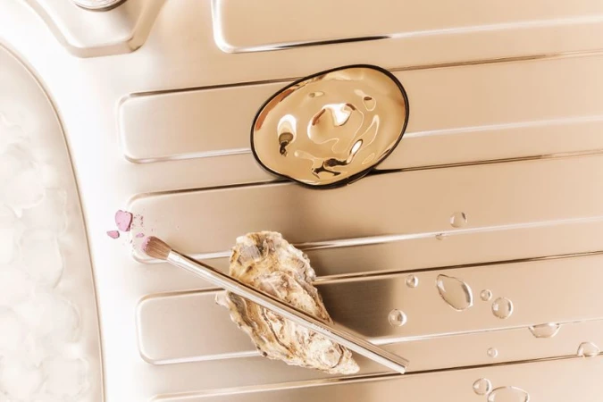 Still life shot of makeup brush, oyster shell, gold dish, water droplets, and pink powder arranged on a textured metallic background.
