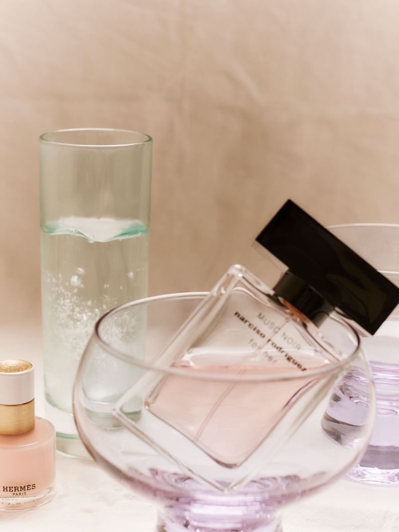 Narciso Rodriguez Musc Noir perfume bottle resting tilted inside a clear glass coupe beside a tall glass of sparkling water.