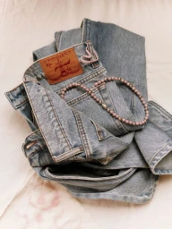 Folded light blue denim jeans featuring a brown leather patch and a beaded necklace draped over the waistband.