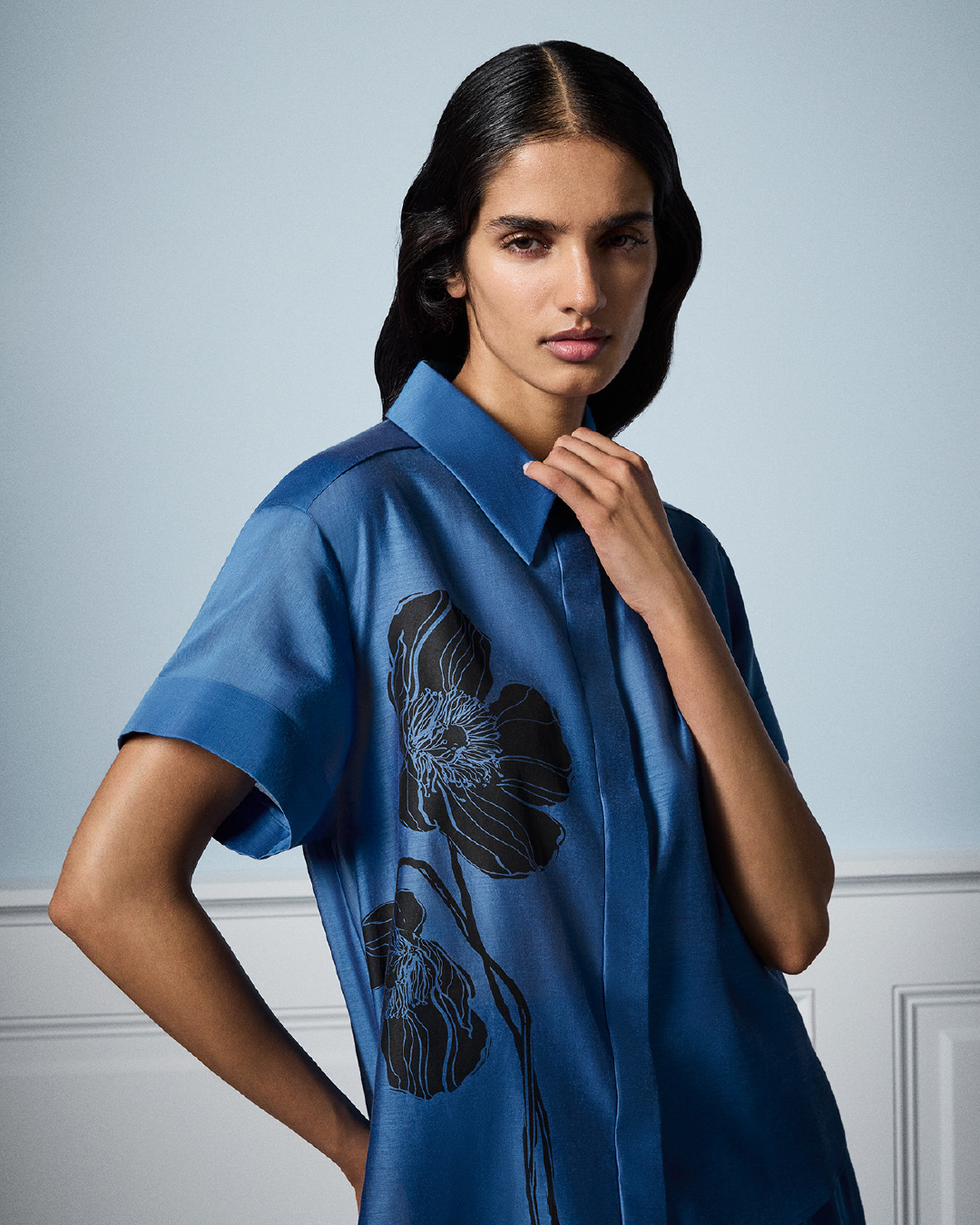 Brunette woman poses in a blue, collared, short-sleeved dress with black flower graphics against a pale blue background.