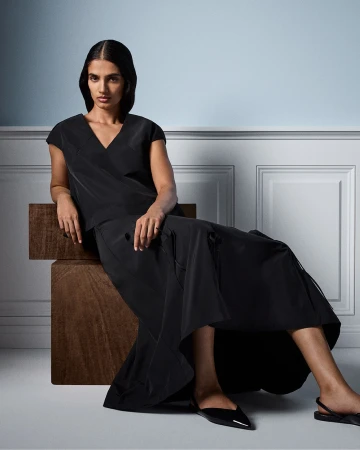An adult female model in a draped black dress and black shoes poses indoors, seated on wooden blocks against a light blue wall.