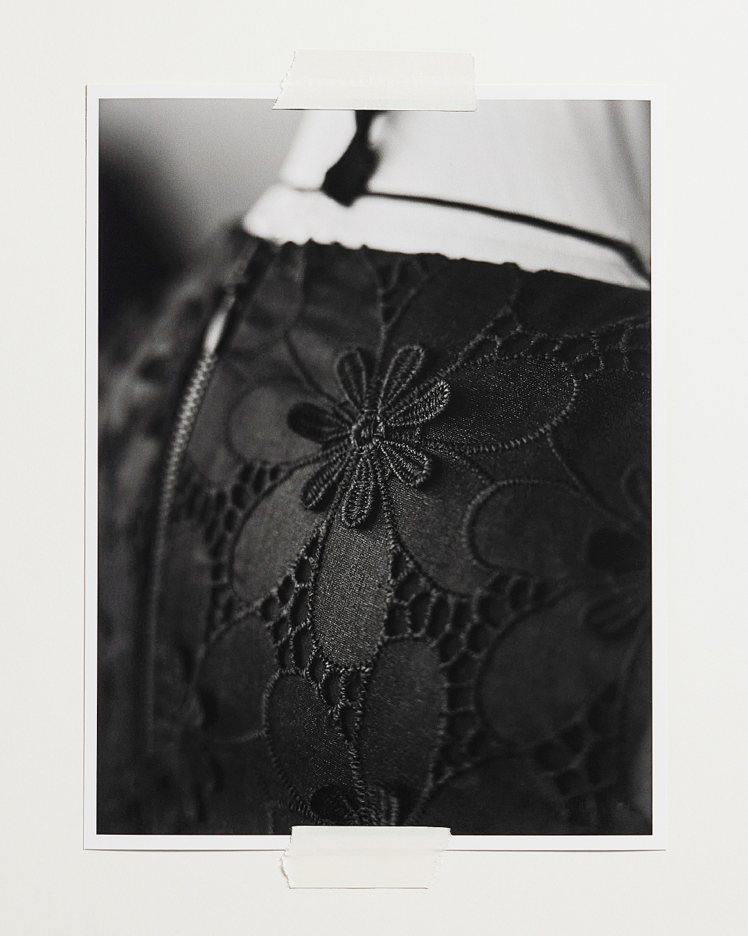 Monochrome close-up of a black lace garment with floral embroidery details, taped to a white wall for a textured, artistic aesthetic.