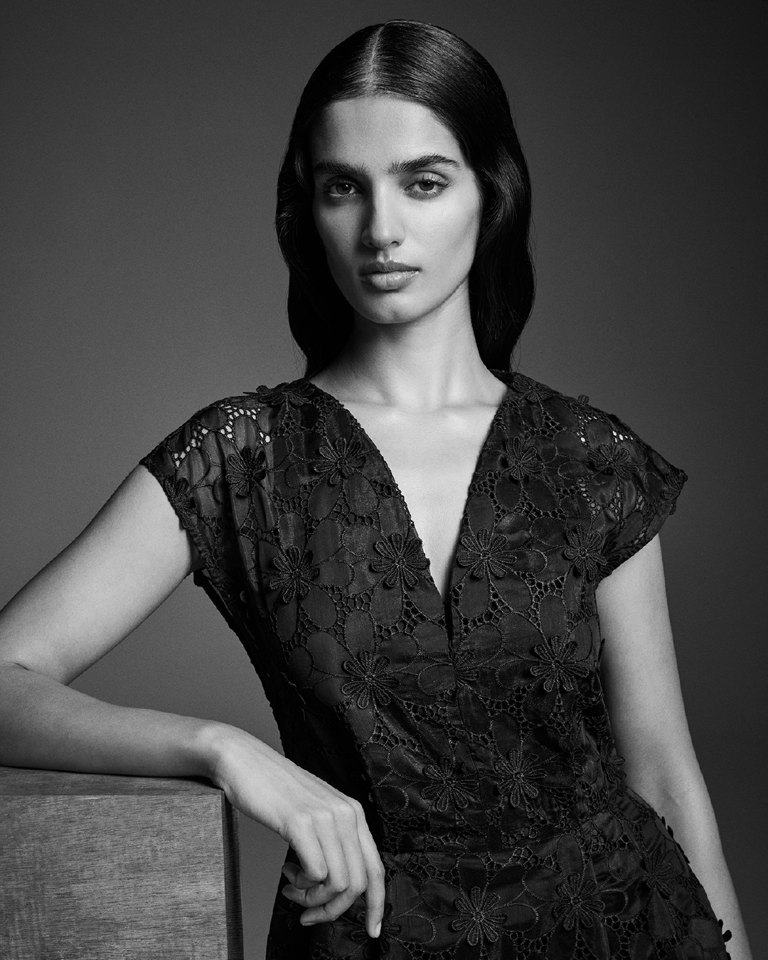 Close-up black and white portrait of a woman in a patterned black lace dress, leaning on a wooden cube with grey background.