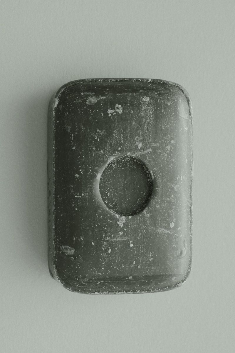 Rectangular gray soap bar with a circular indentation in the center, casting a soft shadow on a plain, light-colored surface.