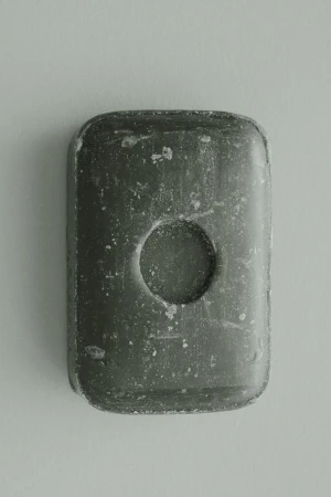 Rectangular gray soap bar with a circular indentation in the center, casting a soft shadow on a plain, light-colored surface.
