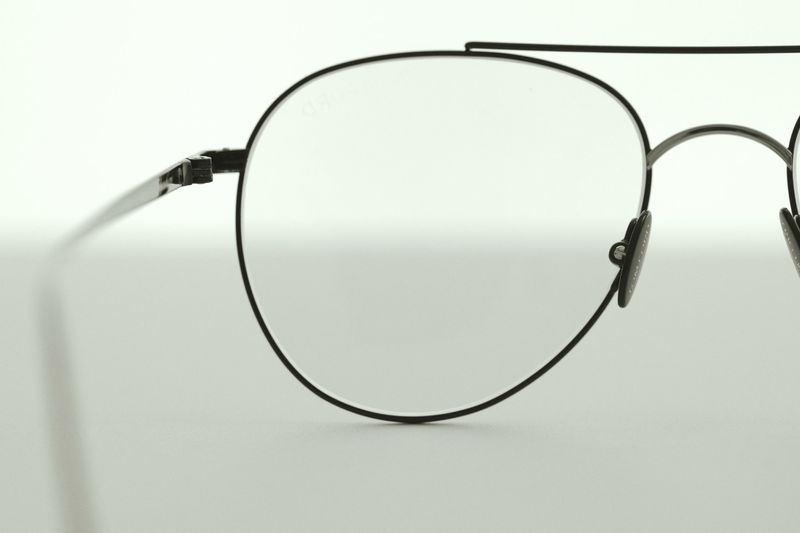 Close-up product shot of black metal aviator glasses with clear lenses against a white background, showcasing their thin frames and design.