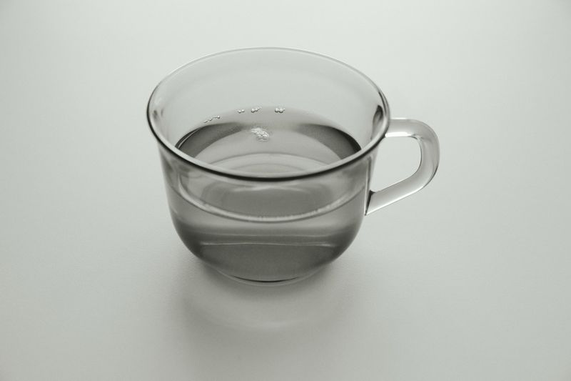 Clear glass mug filled with liquid. The mug is on a white surface with neutral lighting. The drink is transparent and still.