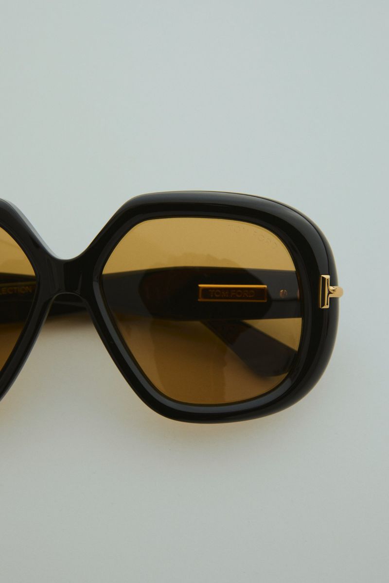 Close-up of glossy black sunglasses featuring square frames and golden brown tinted lenses, set against a neutral light background.