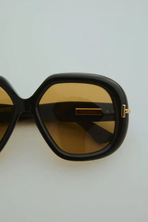 Close-up of glossy black sunglasses featuring square frames and golden brown tinted lenses, set against a neutral light background.