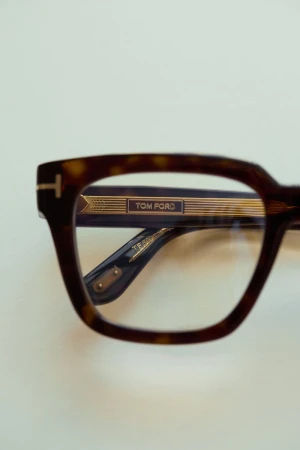 Close-up of tortoiseshell Tom Ford eyeglasses on a white surface, highlighting the frame's pattern, gold accents, and lens shape.