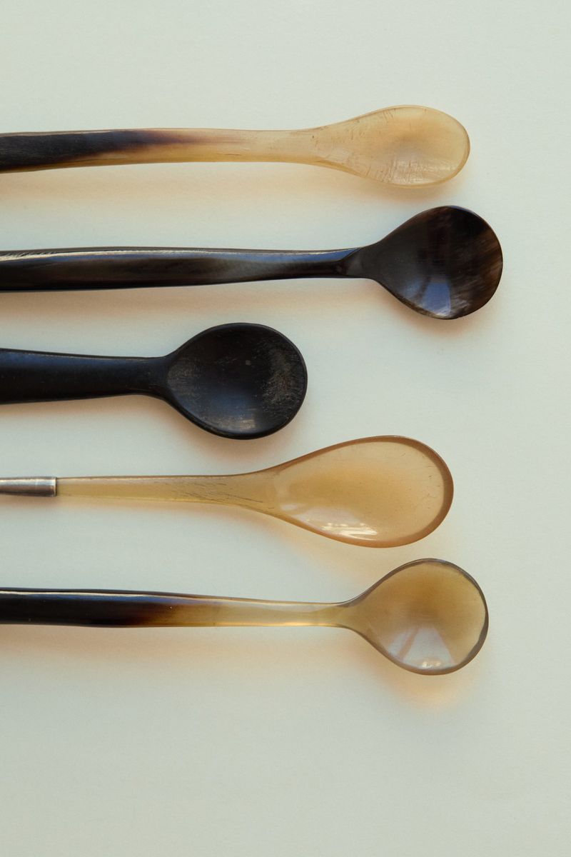 Five spoons of varying colors and materials are arranged in a row on a plain white surface in this top-down studio shot.
