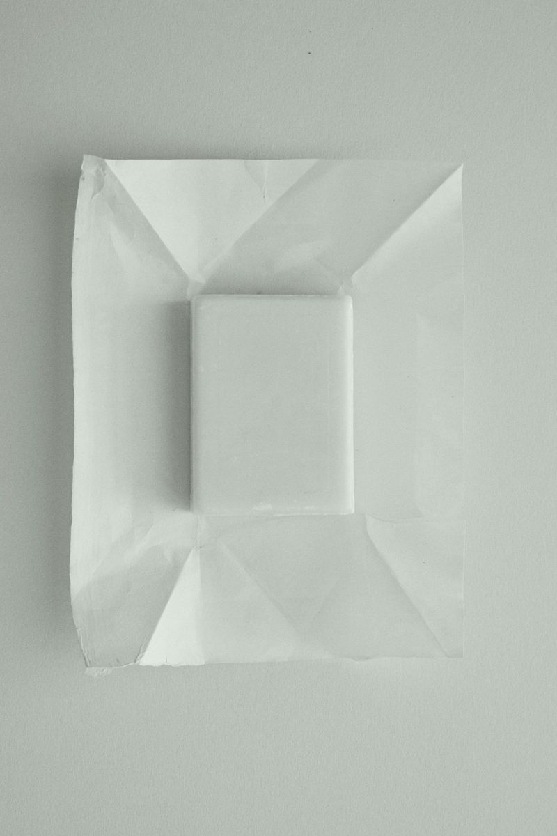 Minimalist composition with a white cube resting on folded white paper, set against an off-white background. Focus on texture and geometry.