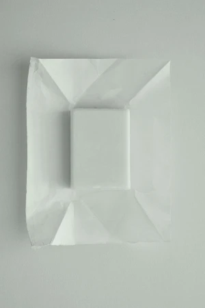 Minimalist composition with a white cube resting on folded white paper, set against an off-white background. Focus on texture and geometry.
