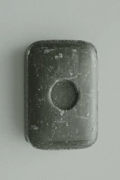 Rectangular dark charcoal bar of soap with a circular central indentation set against a smooth pale grey seamless background.