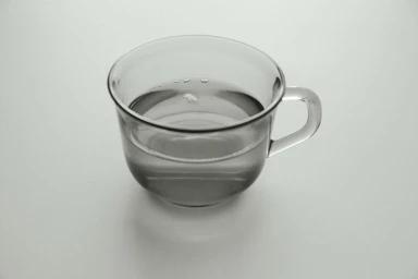 A clear glass cup filled with water casting soft shadows on a smooth, neutral light grey seamless background.