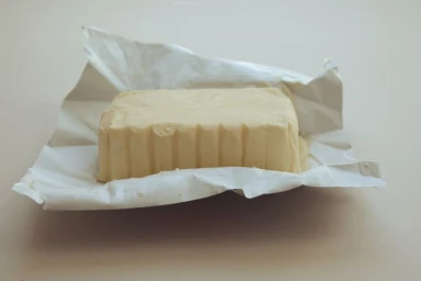 A rectangular block of pale yellow butter rests unwrapped on creased white and silver paper packaging on a neutral surface.