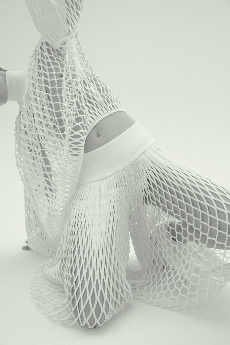 Monochrome fashion shot of a slender adult female model kneeling, posing in a semi-sheer white net outfit on a bright neutral background.