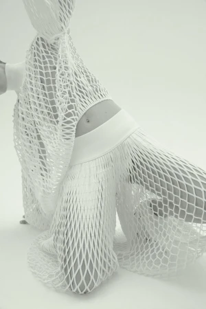 Monochrome fashion shot of a slender adult female model kneeling, posing in a semi-sheer white net outfit on a bright neutral background.