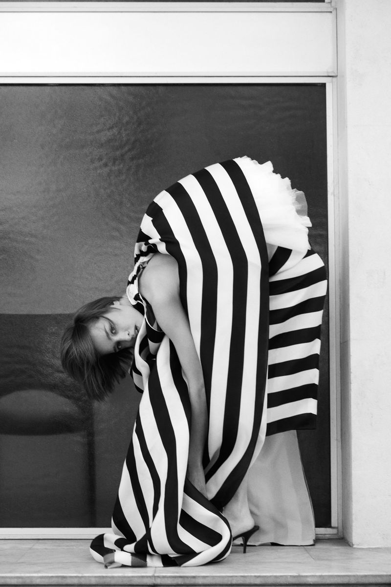 Monochrome image of a woman in a striped dress bending over in front of a window and white wall, with a high-fashion editorial aesthetic.