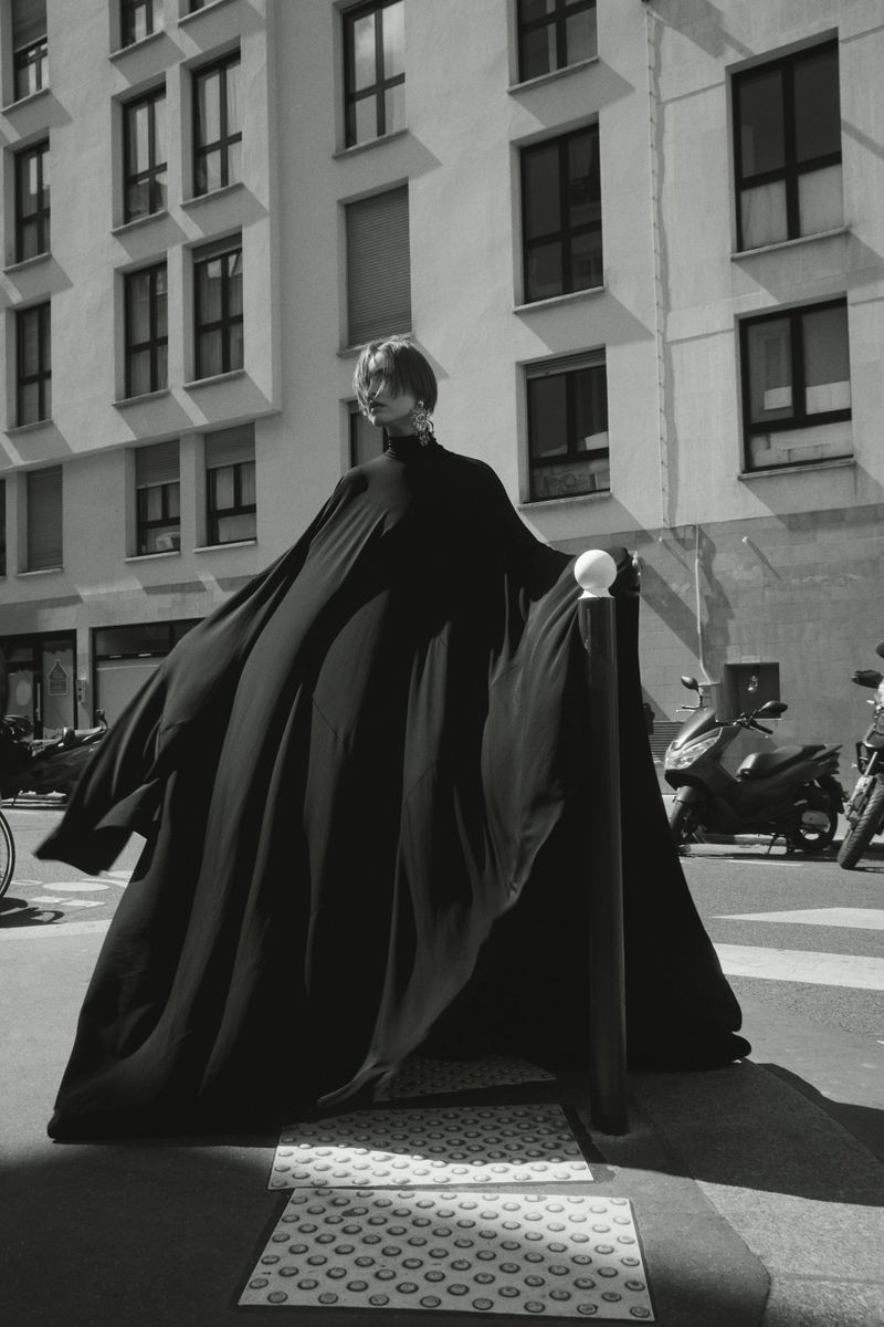 Monochromatic fashion portrait of a model in a black gown standing on a city street, building and scooter in the background.