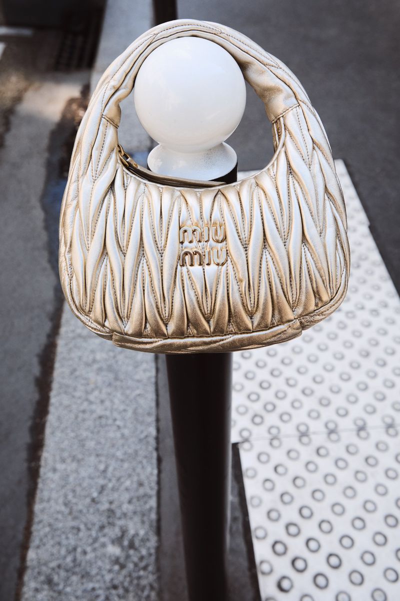 Beige Miu Miu Matelassé handbag with a quilted finish, handle, and a visible Miu Miu logo, resting on a white and black bollard outdoors.