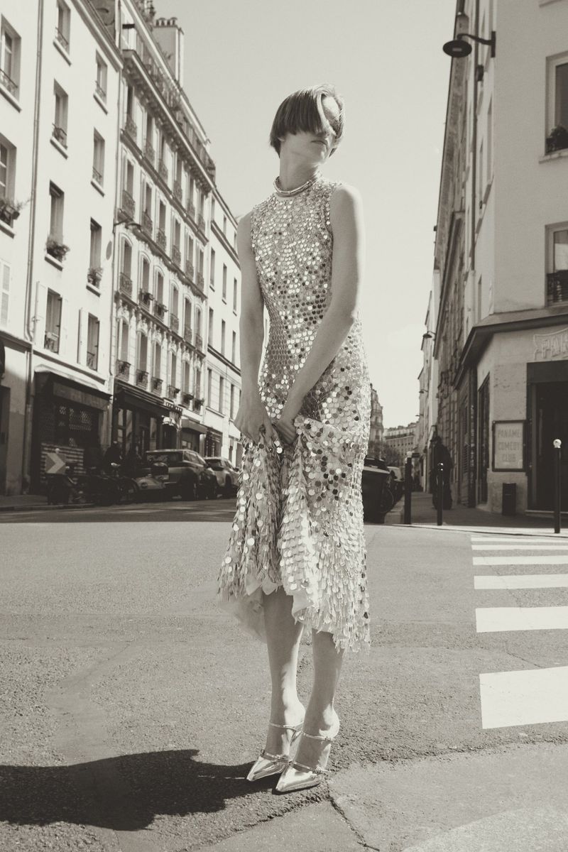 A woman in a shimmering dress and heels poses on a crosswalk in a black-and-white urban scene in Paris, France, buildings in background.
