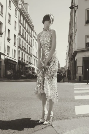 A woman in a shimmering dress and heels poses on a crosswalk in a black-and-white urban scene in Paris, France, buildings in background.