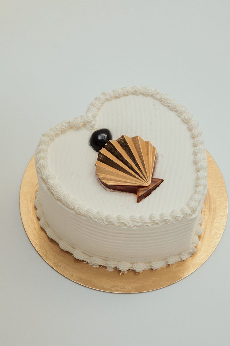 Heart-shaped cake on gold base, decorated with white frosting, chocolate seashell and chocolate ball. White background.