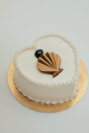 Heart-shaped cake on gold base, decorated with white frosting, chocolate seashell and chocolate ball. White background.
