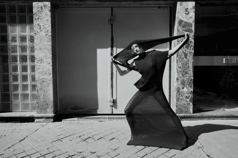 Monochrome fashion shot of a woman in a pleated dress and headpiece posing against a building with doors and glass block windows.