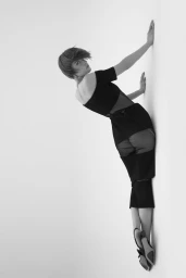 Model Lea Lukek wearing a black sheer-paneled midi dress leaning against a white studio wall with hands outstretched.