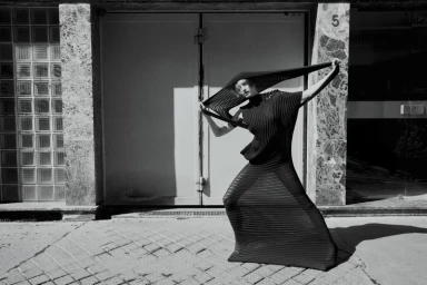 Model Lea Lukek poses outdoors in an avant-garde black pleated dress holding a wide sculptural hood overhead in direct sunlight.
