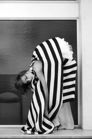 Model Lea Lukek bends forward draped in a bold black and white striped garment beside a large dark window.