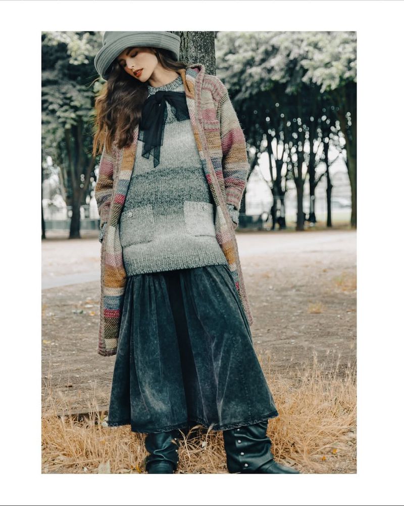A woman stands in an outdoor setting, modelling a gray knit sweater, long gray skirt, a hat, black boots and a multicolor striped coat.