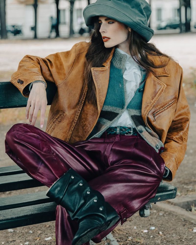Woman in a hat and leather jacket poses on a park bench, wearing maroon pants and black boots, with a park and trees in the background.