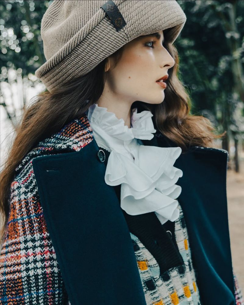 Brunette woman in profile wearing a beige hat and colorful patterned coat with white ruffle collar standing in an outdoor setting.
