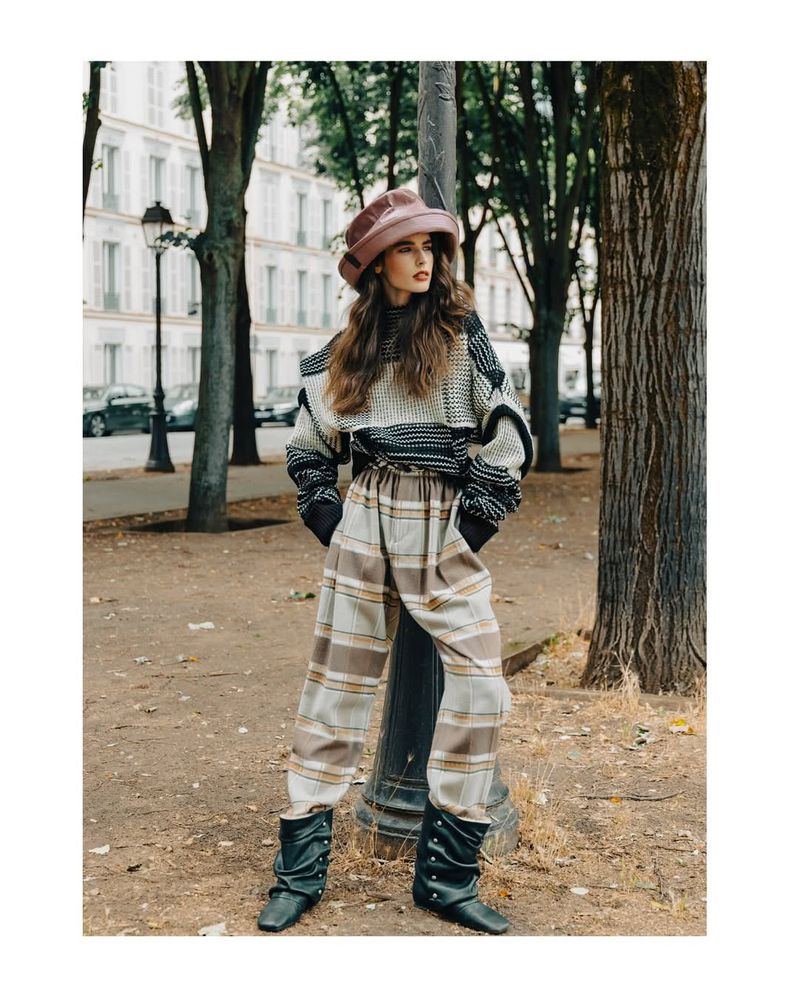 A fashionable adult woman in a hat, patterned sweater, and pants poses in an outdoor urban environment with parked cars.