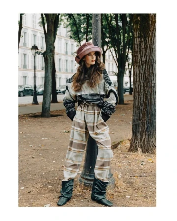 A fashionable adult woman in a hat, patterned sweater, and pants poses in an outdoor urban environment with parked cars.