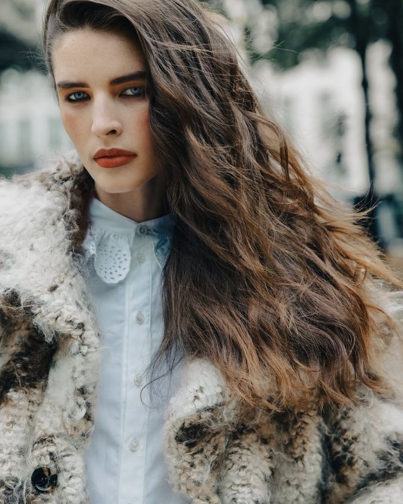Close-up portrait of a young woman with long, wavy brown hair, wearing a light blue collared shirt and textured coat in an outdoor setting.