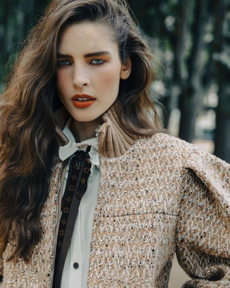 A fashionable woman poses in a tweed jacket, with long wavy brown hair, bold makeup, and an outdoor backdrop of trees and greenery.